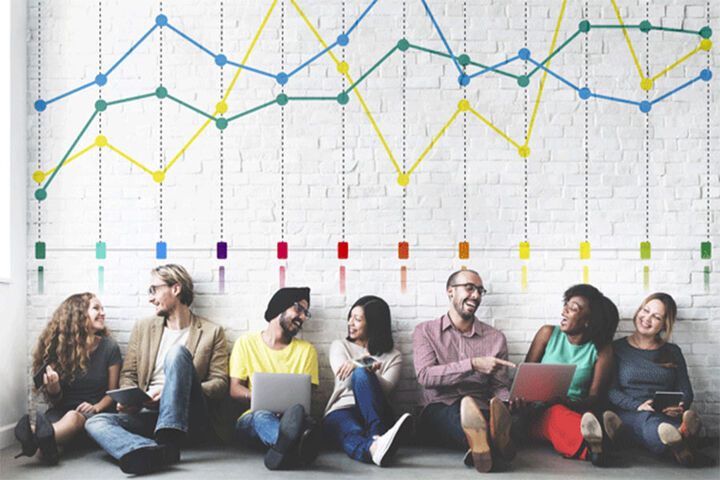 A group of students with laptops sits against a wall that has a chart painted on it.